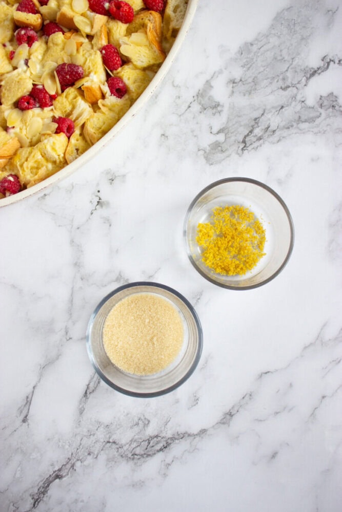 A baking dish with bread pudding and berries is on a marble surface next to two small bowls containing lemon zest and granulated sugar.