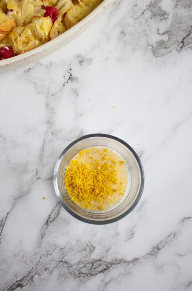 A glass bowl with yellow lemon zest and sugar sits on a marble countertop next to a partially visible baking dish.