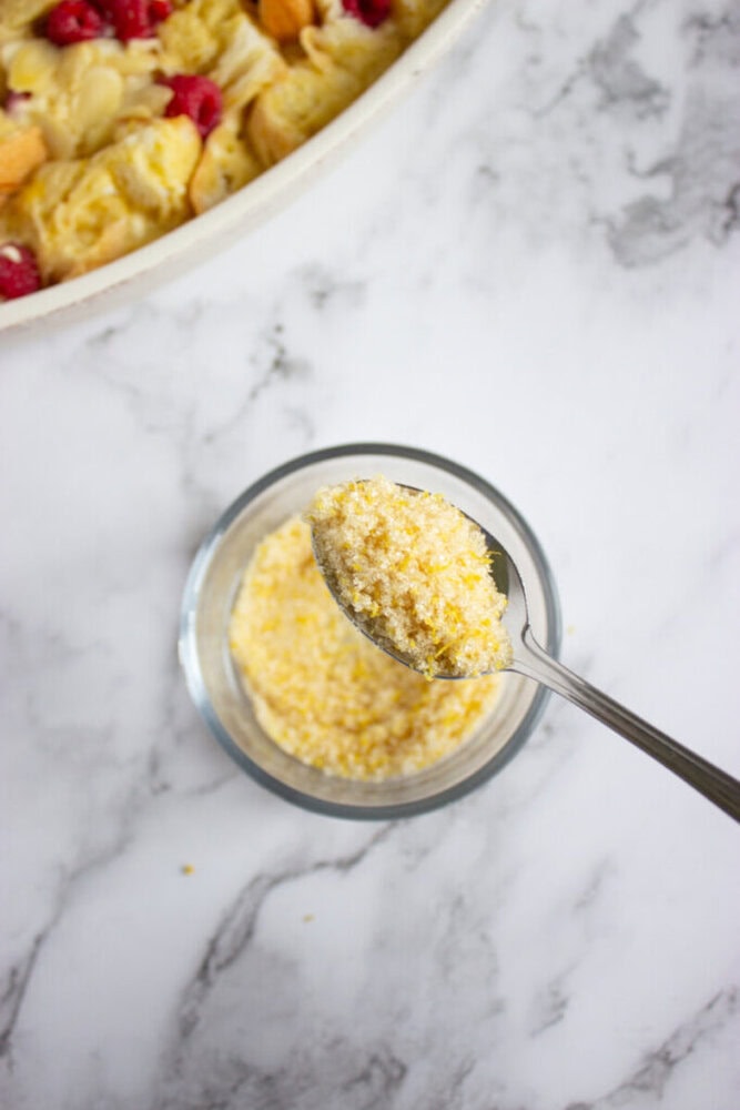 A spoon holding a yellow crumbly mixture over a small glass bowl, with part of a baking dish visible in the top left corner on a marble surface.