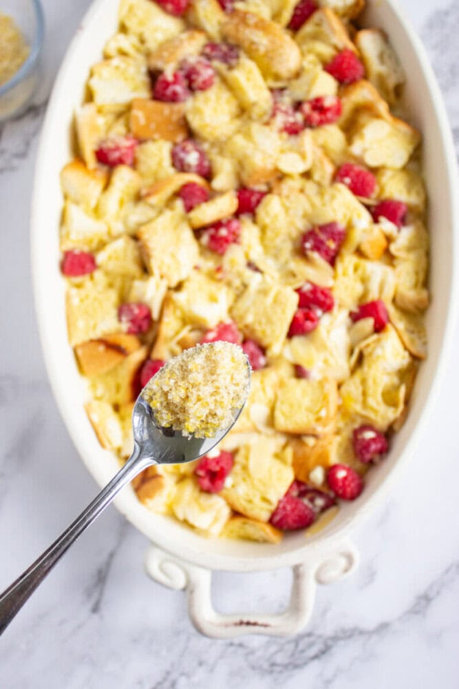 A spoonful of coarse sugar is held above a baking dish filled with pieces of bread, raspberries, and a custard mixture, ready to be baked.