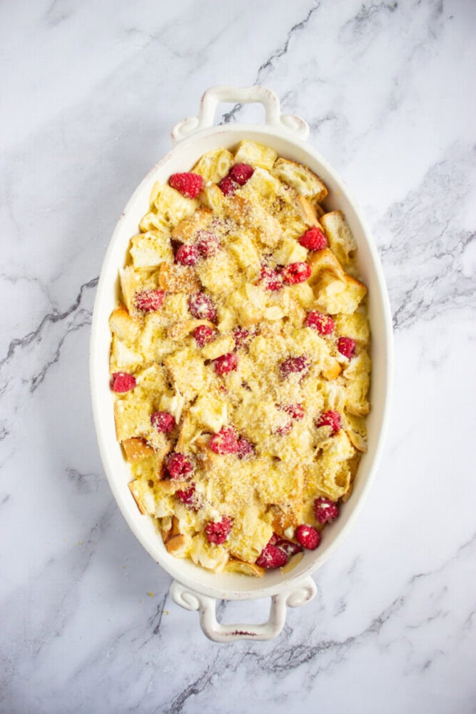 An oval white baking dish filled with unbaked bread pudding, topped with fresh raspberries and a crumbly mixture, on a marble surface.