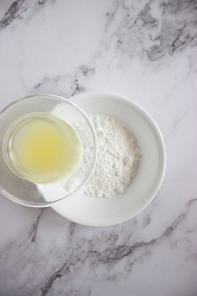 A bowl of powdered sugar next to a glass jar containing lemon juice on a white marble surface.