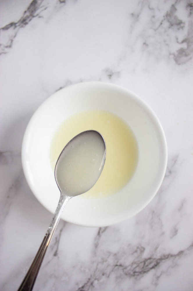 A metal spoon holds a cloudy liquid above a white bowl containing more of the same liquid, set on a white marble surface.