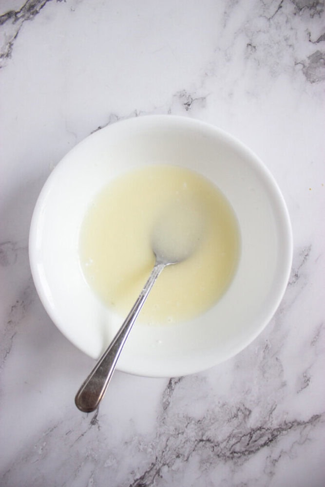 A white bowl containing a pale yellow liquid with a metal spoon, placed on a white marble surface.