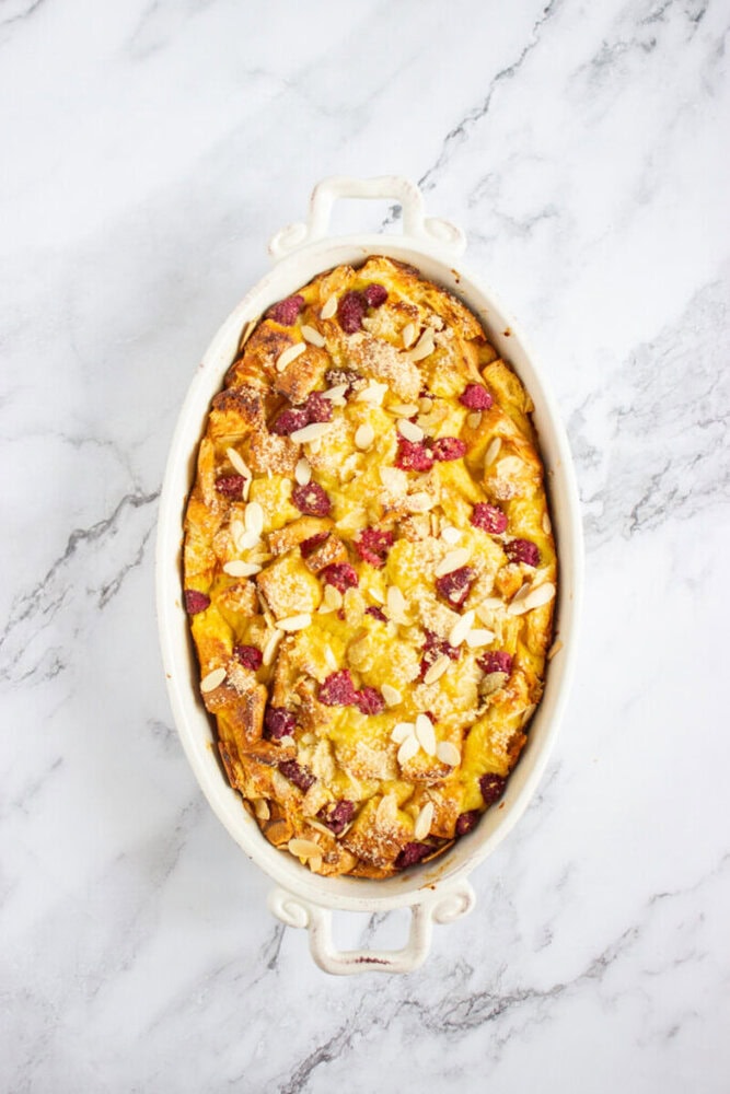 An oval baking dish filled with bread pudding topped with sliced almonds and raspberries, set on a marble surface.