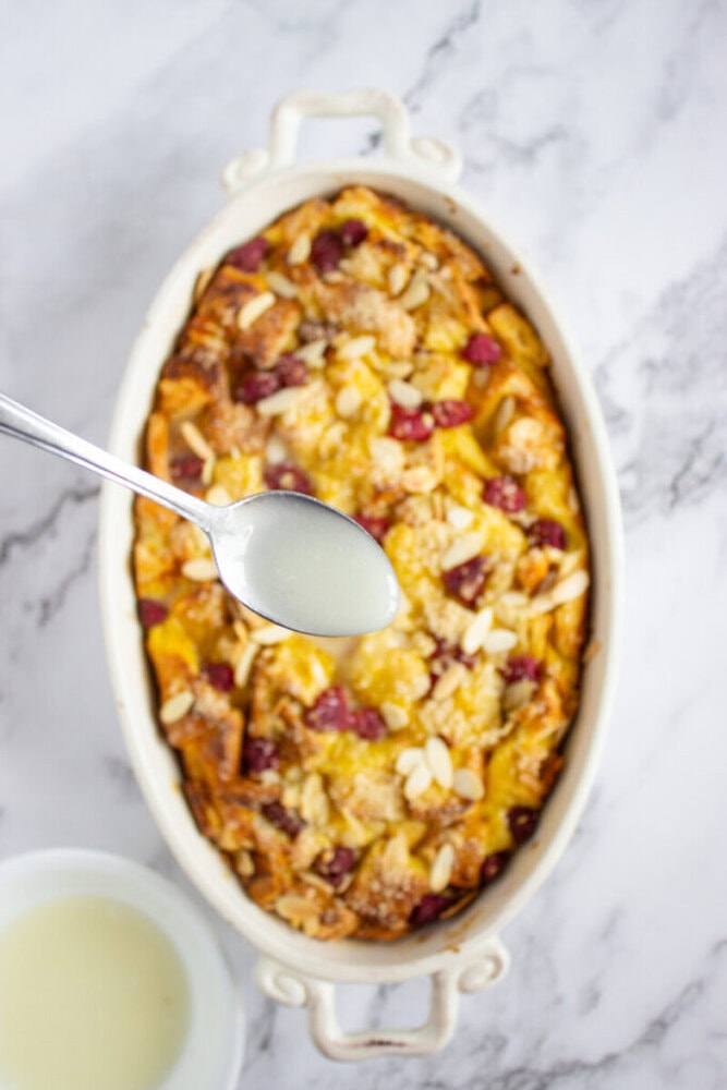 A spoon holding glaze is positioned above a baked fruit and nut casserole in an oval dish, with a bowl of glaze nearby on a marble surface.