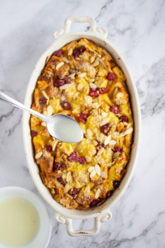 A spoon holds a white liquid above a baked casserole topped with sliced almonds and berries, placed in an oval dish on a marble surface.