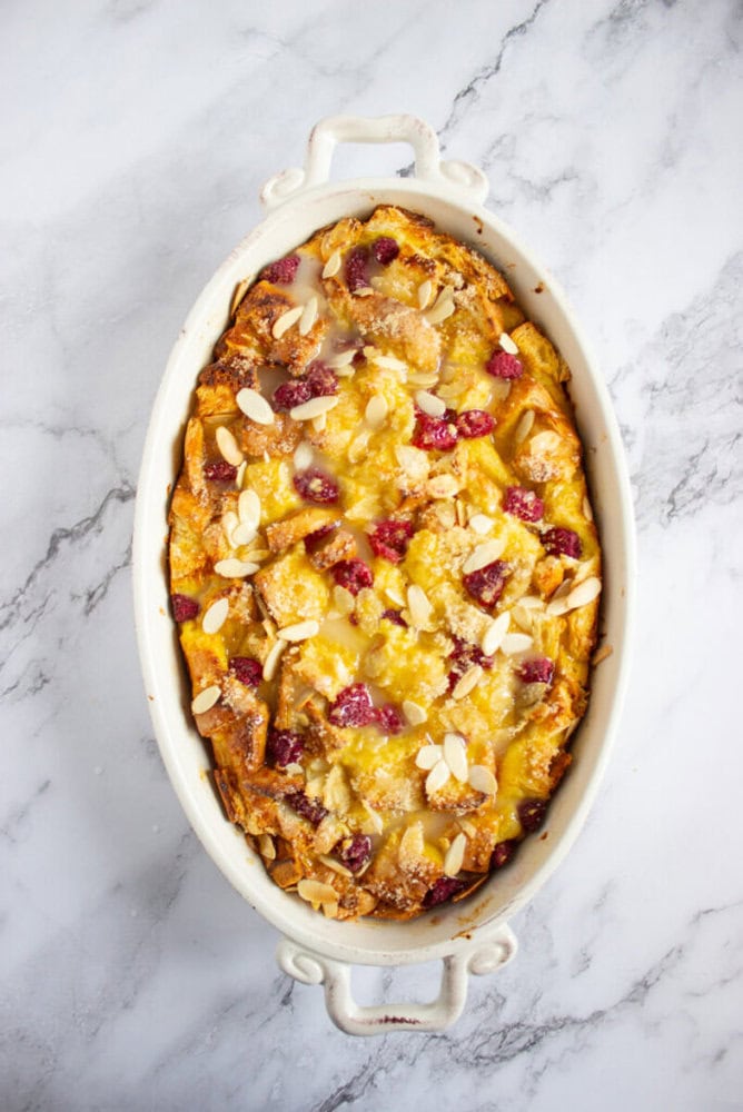 A baked bread pudding in an oval white dish, topped with sliced almonds and raspberries, on a marble surface.