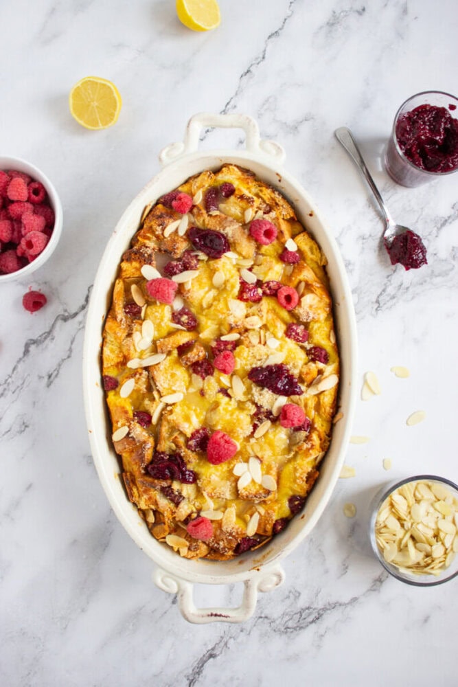 An oval baking dish filled with bread pudding topped with raspberries, jam, and sliced almonds sits on a marble surface, surrounded by extra ingredients.