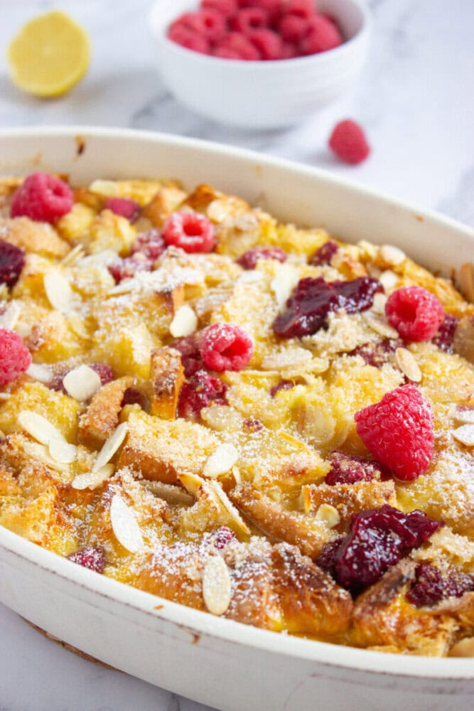 A baked bread pudding topped with fresh raspberries, jam, sliced almonds, and powdered sugar in a white dish; a bowl of raspberries and a lemon are in the background.