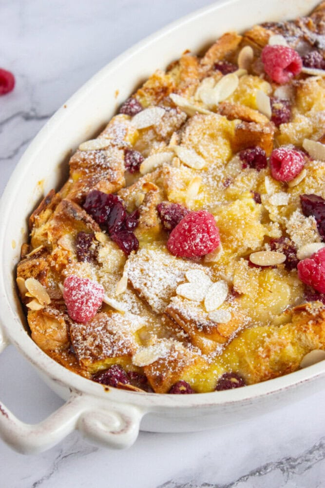 Baked bread pudding in a white dish, topped with sliced almonds, raspberries, dried cranberries, and powdered sugar.