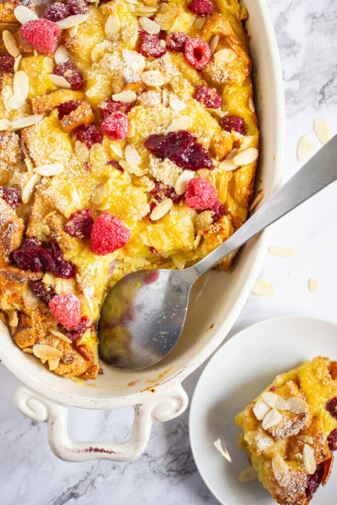 Baked bread pudding topped with raspberries, almond slices, and powdered sugar in a white dish, with a serving spoon and a portion on a plate nearby.