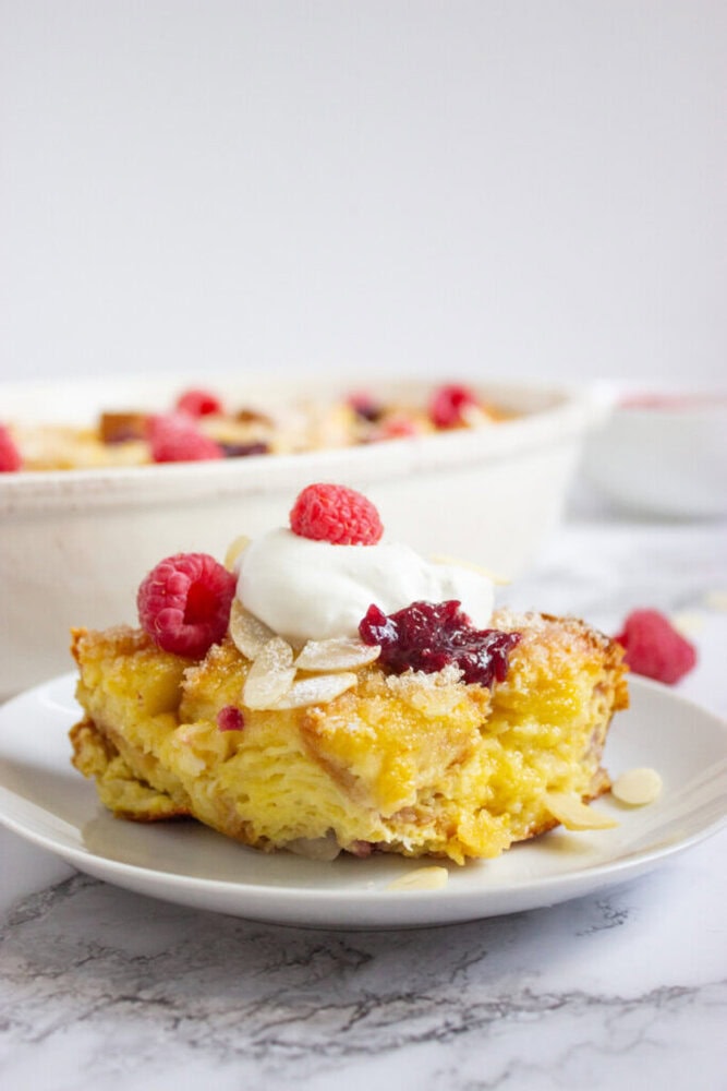 A slice of bread pudding topped with whipped cream, raspberry jam, fresh raspberries, and sliced almonds on a white plate, with a baking dish in the background.