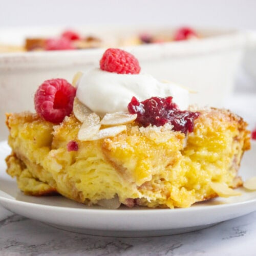 A slice of bread pudding topped with whipped cream, raspberry sauce, fresh raspberries, almond slices, and a dusting of powdered sugar on a white plate.