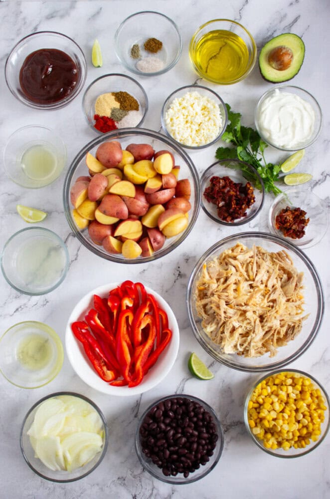 Various ingredients in glass bowls on a marble surface, including chopped potatoes, shredded chicken, black beans, corn, red bell pepper, onion, cheese, spices, sauces, and herbs.