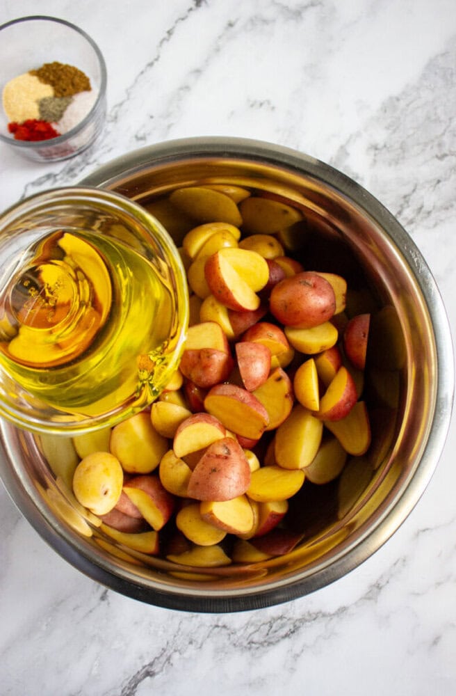 A metal bowl filled with halved red potatoes sits on a marble surface; olive oil is being poured over them. A small bowl of spices is visible nearby.