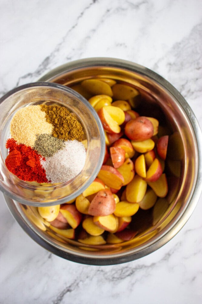 A metal bowl of halved red potatoes with a small glass bowl of assorted spices, including paprika, salt, pepper, and garlic powder, placed above it on a marble surface.