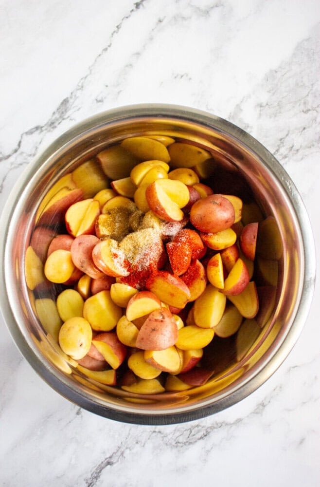 Halved red and yellow baby potatoes with various seasonings in a stainless steel mixing bowl on a marble countertop.