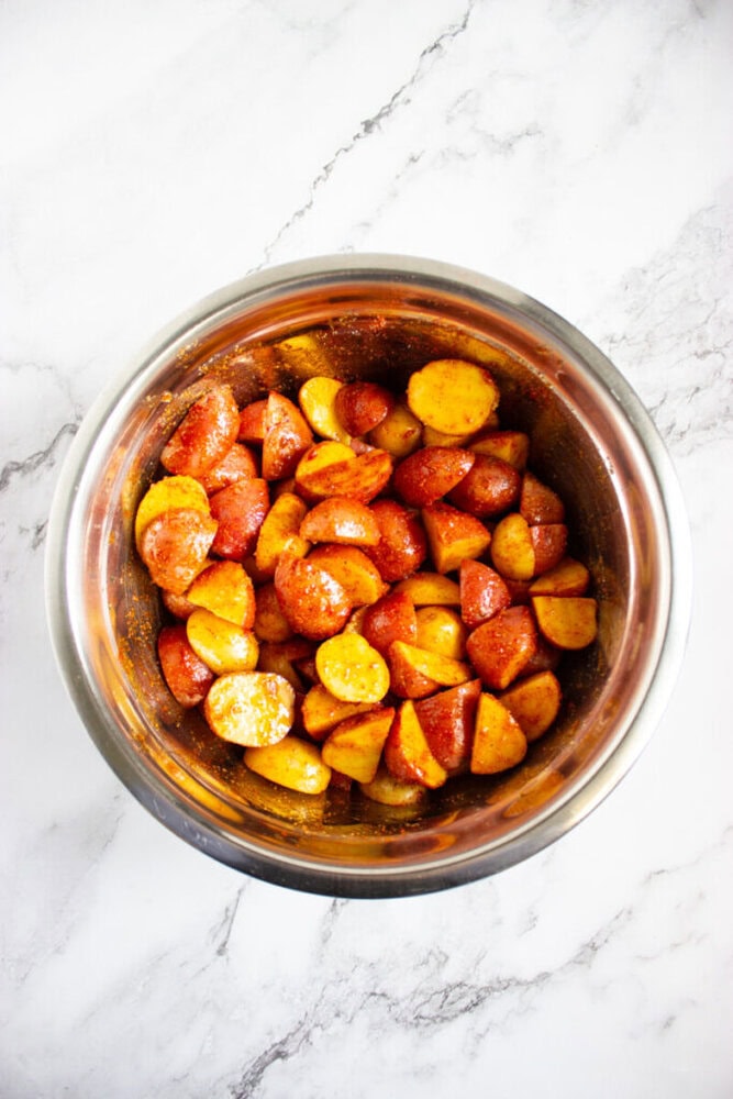 A stainless steel bowl filled with sliced, seasoned red and yellow baby potatoes on a white marble surface.