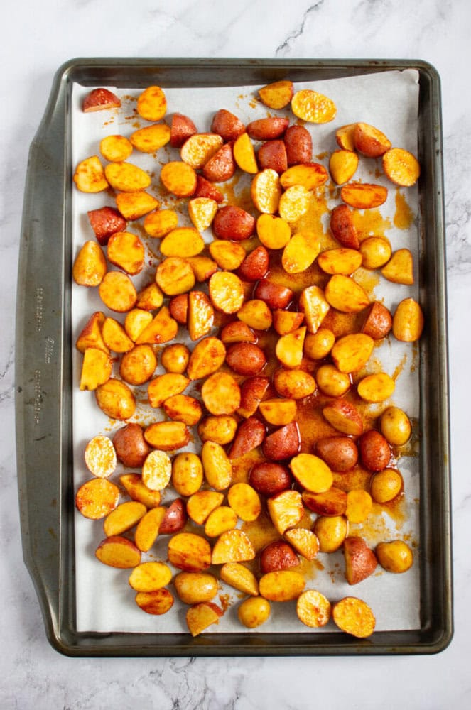 A baking sheet lined with parchment paper holds halved, seasoned red potatoes ready for roasting on a marble countertop.