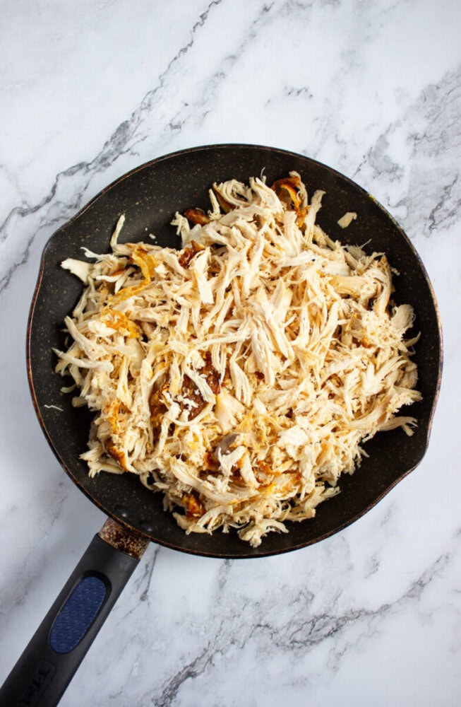A black frying pan filled with shredded cooked chicken sits on a white marble countertop.