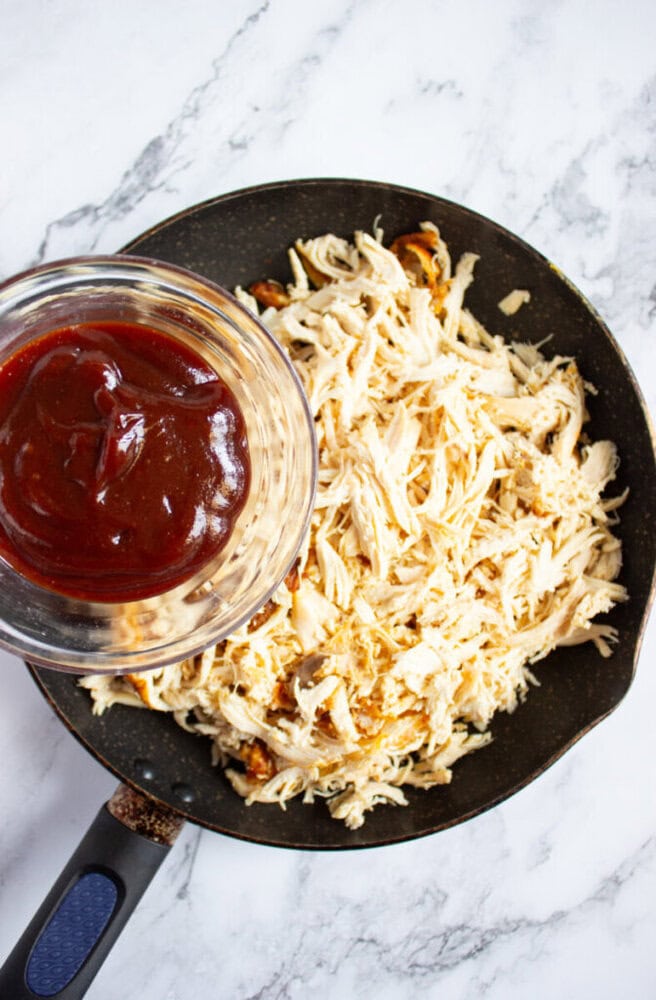 A skillet of shredded chicken next to a glass bowl of barbecue sauce on a marble countertop.