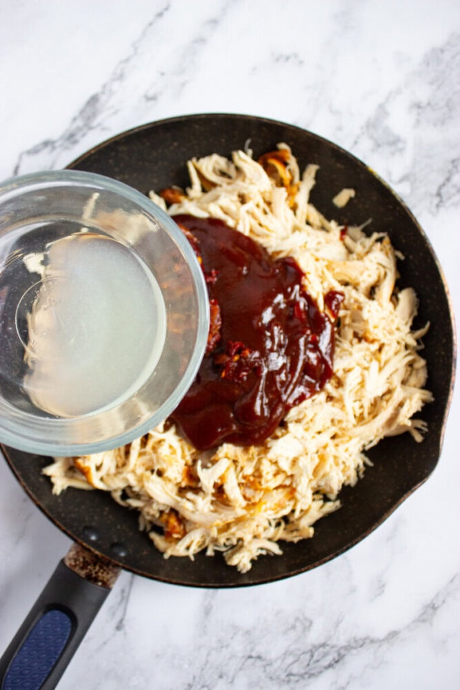 A skillet with shredded chicken topped with barbecue sauce, and a hand pouring liquid from a glass onto the mixture, set on a marble surface.