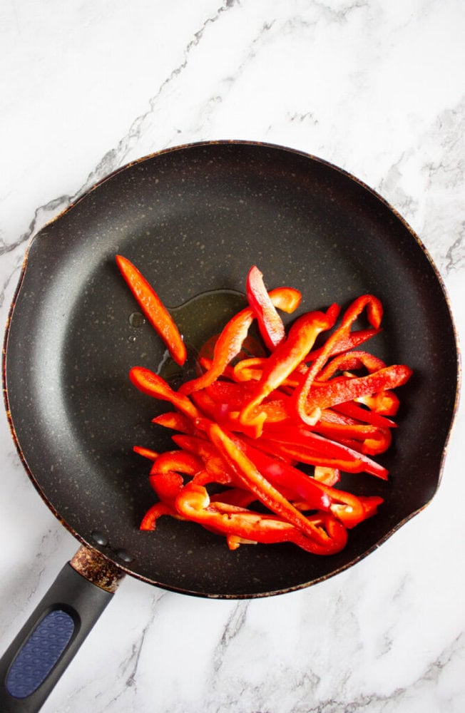 A nonstick frying pan with sliced red bell peppers and a small amount of oil on a marble surface.