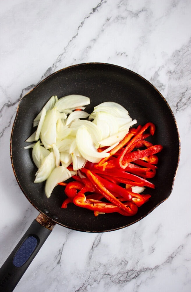 A frying pan with sliced onions and red bell peppers on a marble surface, ready to be cooked.