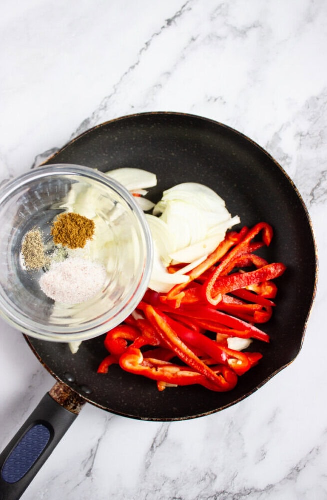A frying pan with sliced red bell peppers and onions next to a small glass bowl containing ground spices on a marble countertop.