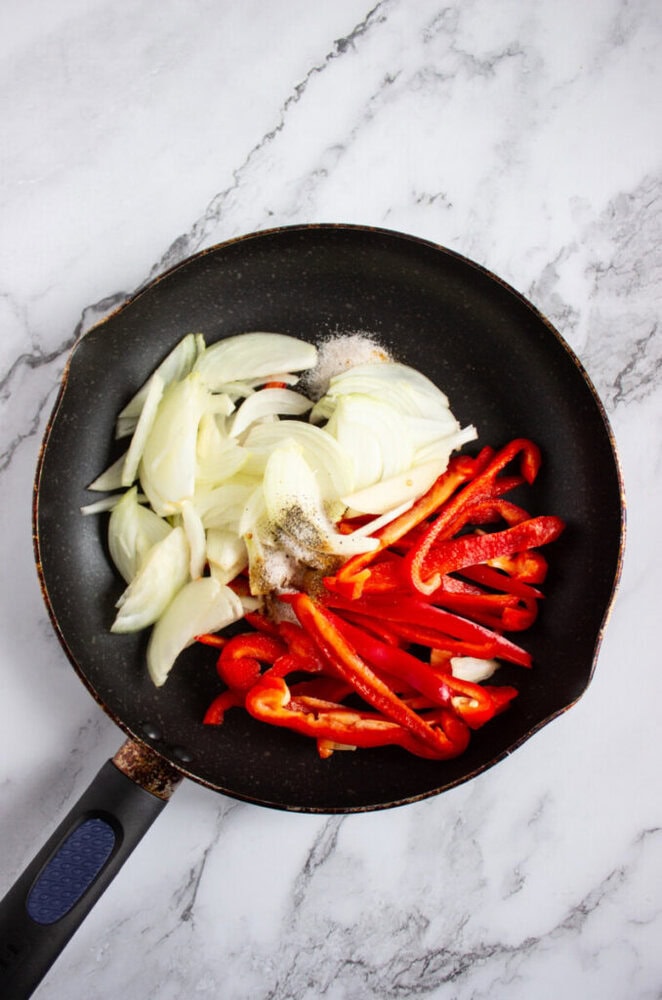 A frying pan with sliced onions and red bell peppers, seasoned with salt and pepper, on a marble countertop.