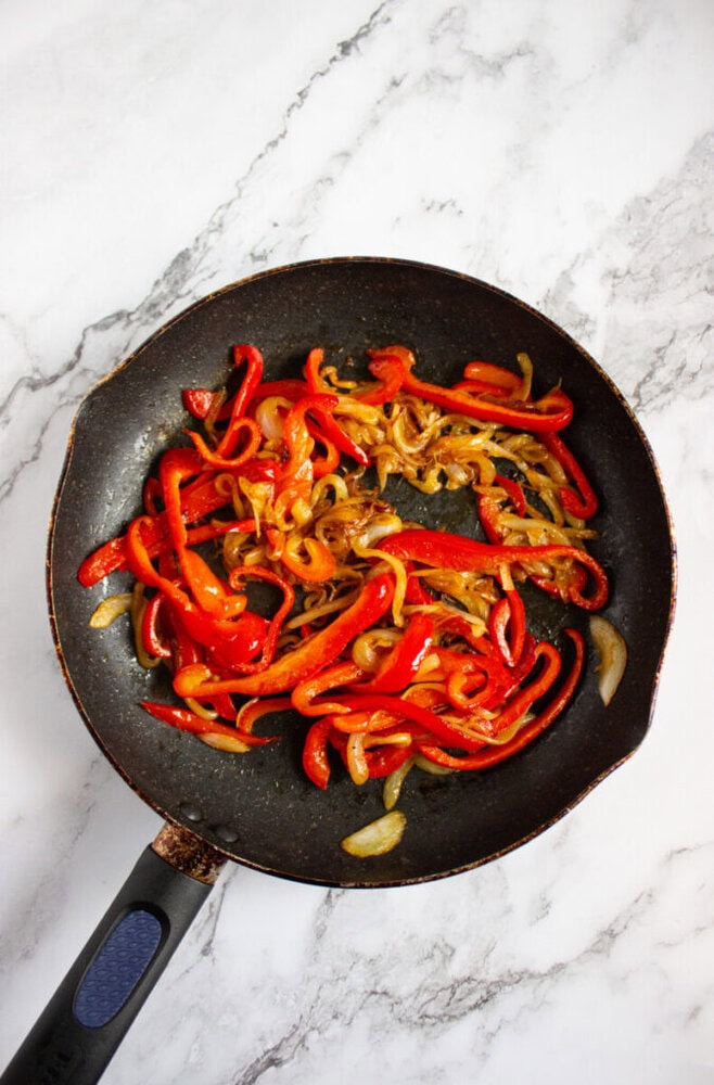 A frying pan on a marble surface containing cooked sliced red bell peppers and onions.