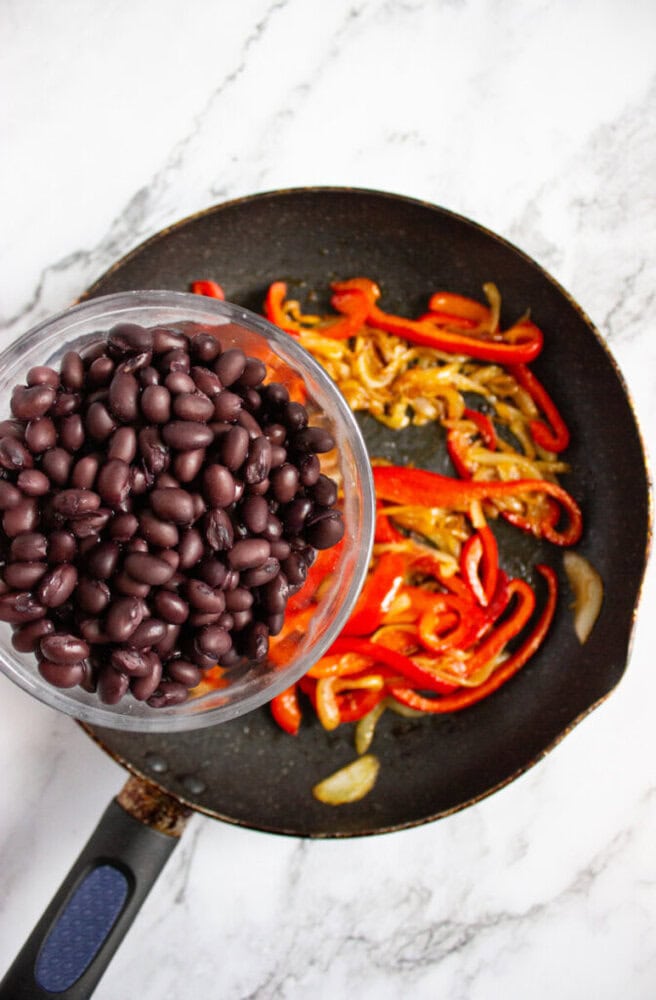 A bowl of black beans held above a frying pan with cooked red bell peppers and onions on a marble countertop.