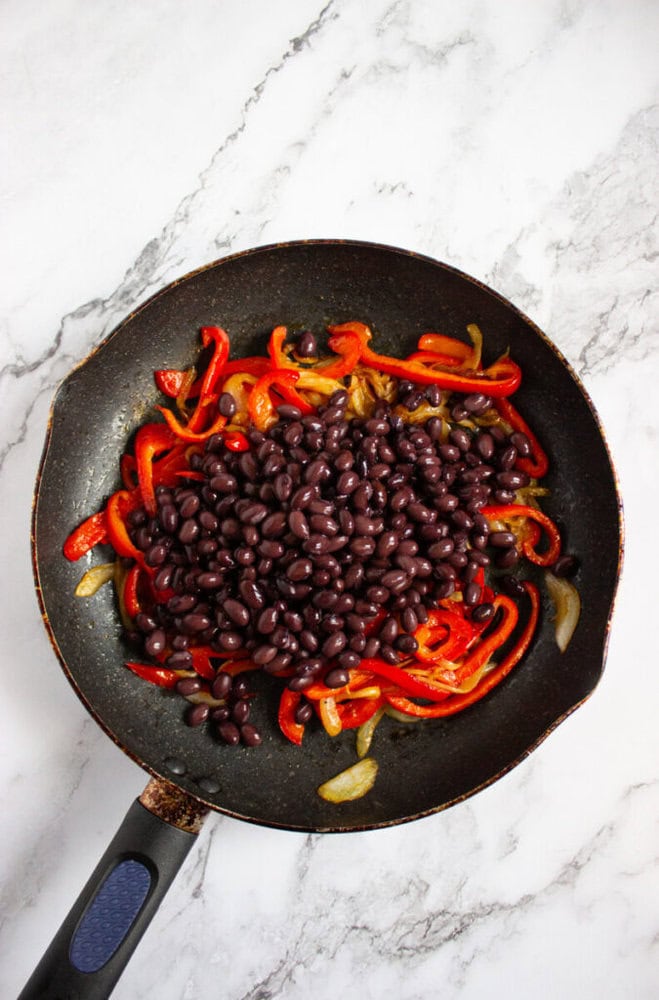A frying pan on a marble surface containing cooked sliced red bell peppers, onions, and a pile of black beans.