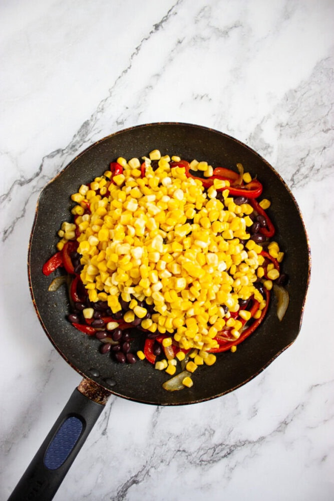A frying pan with corn, black beans, red bell pepper strips, and onions on a white marble surface.