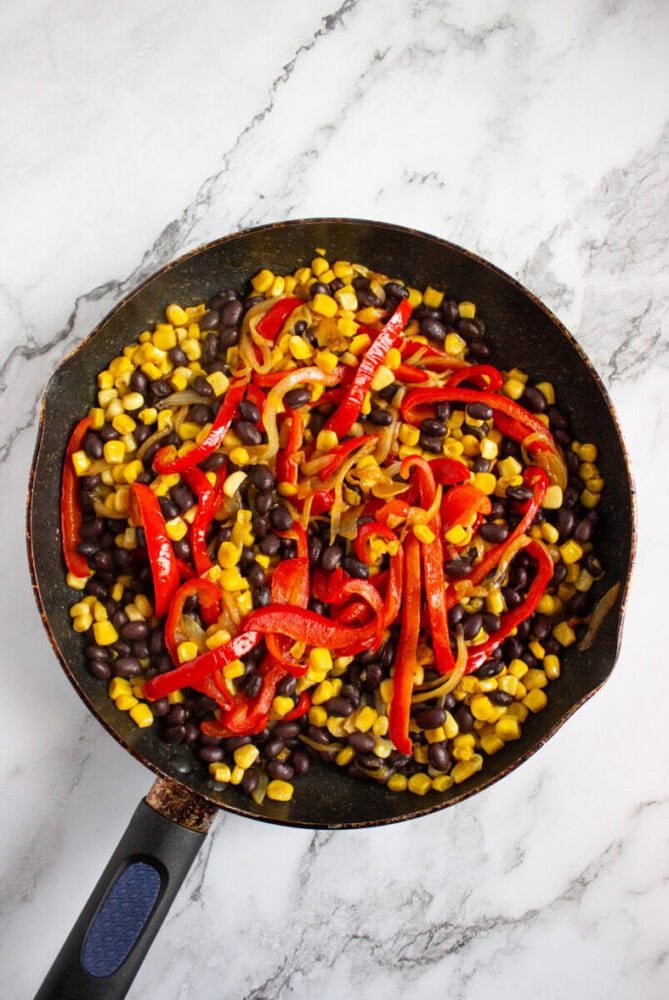 A skillet containing black beans, corn, and sliced red bell peppers sits on a white marble surface.