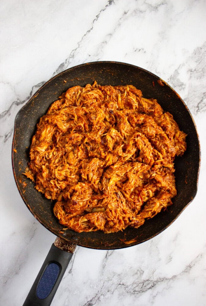 A frying pan filled with shredded, seasoned meat in a reddish sauce, placed on a white marble surface.