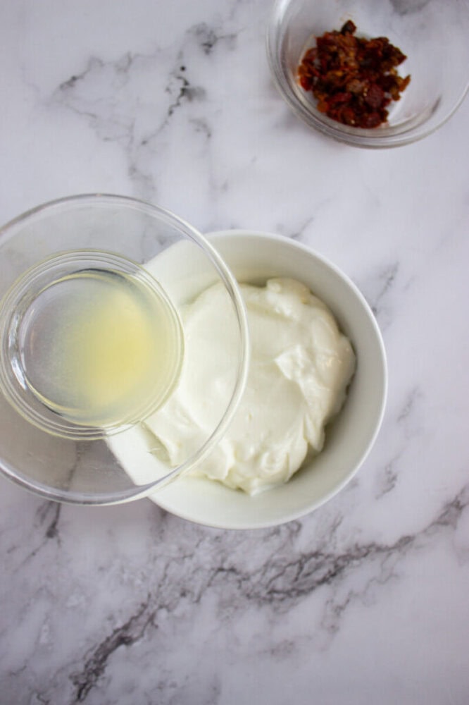 A bowl of plain yogurt is being combined with lemon juice from a small glass bowl; chopped sun-dried tomatoes are in a separate bowl nearby on a marble surface.