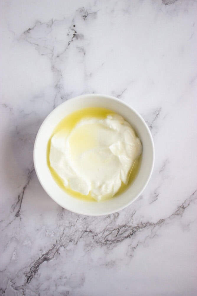 A white bowl filled with plain yogurt topped with a layer of yellow liquid, placed on a white marble surface.