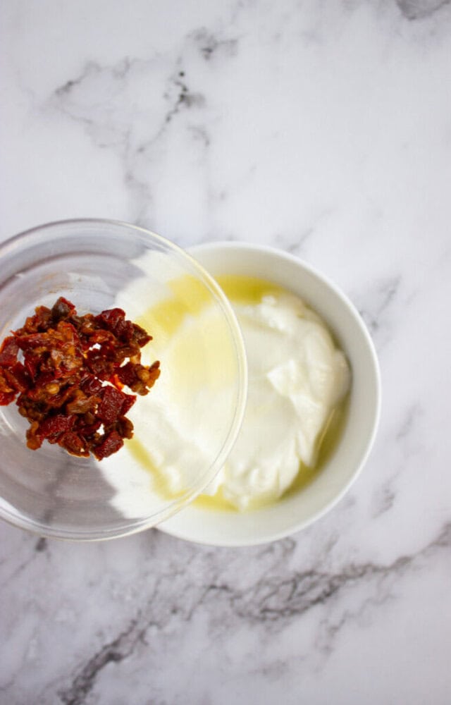 A glass bowl with chopped sun-dried tomatoes is being poured into a white bowl of yogurt with olive oil, on a marble surface.