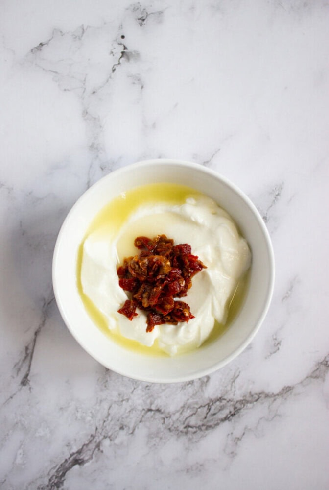 A white bowl containing yogurt topped with chopped sun-dried tomatoes and a drizzle of olive oil, placed on a marble surface.