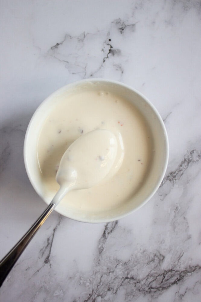 A white bowl filled with creamy white sauce, with a metal spoon resting inside, placed on a light marble surface.