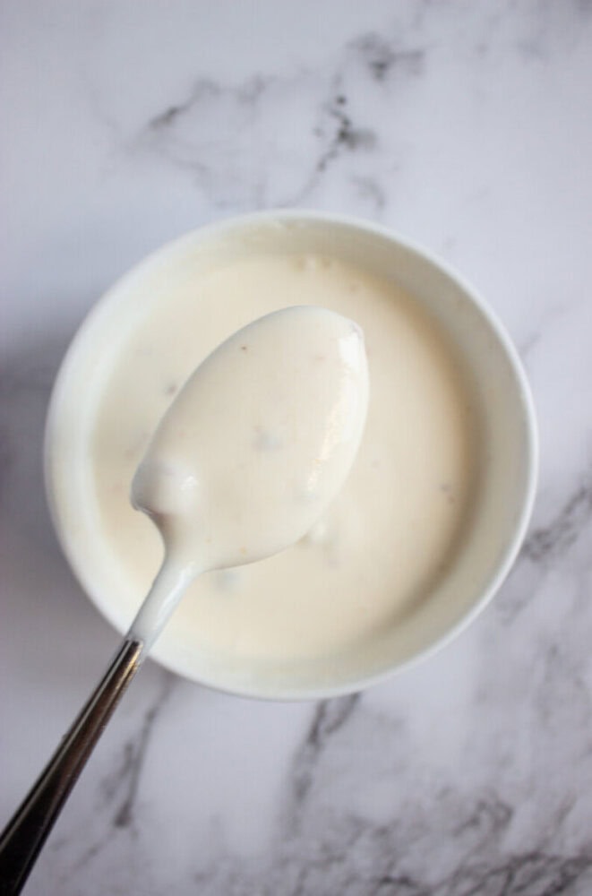A spoonful of creamy white sauce held above a bowl of the same sauce on a marbled surface.