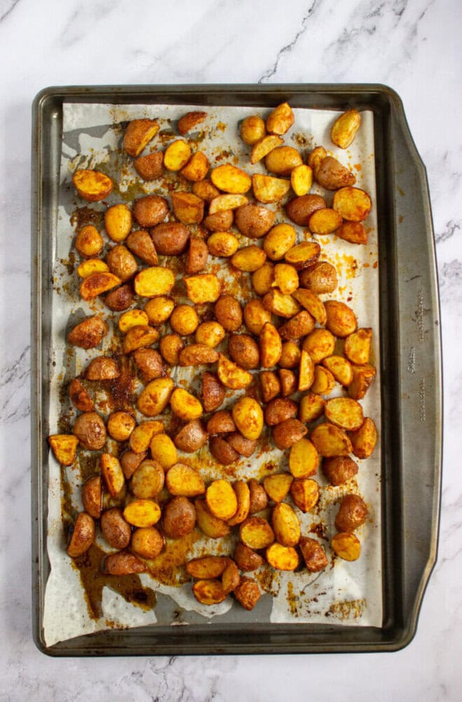 A baking sheet lined with parchment paper holds roasted, seasoned potato pieces on a marble surface.