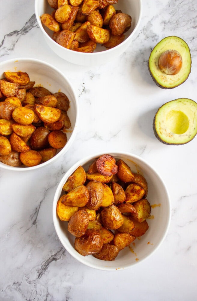 Three white bowls filled with seasoned roasted potatoes are arranged on a marble surface next to a halved avocado with the pit intact.