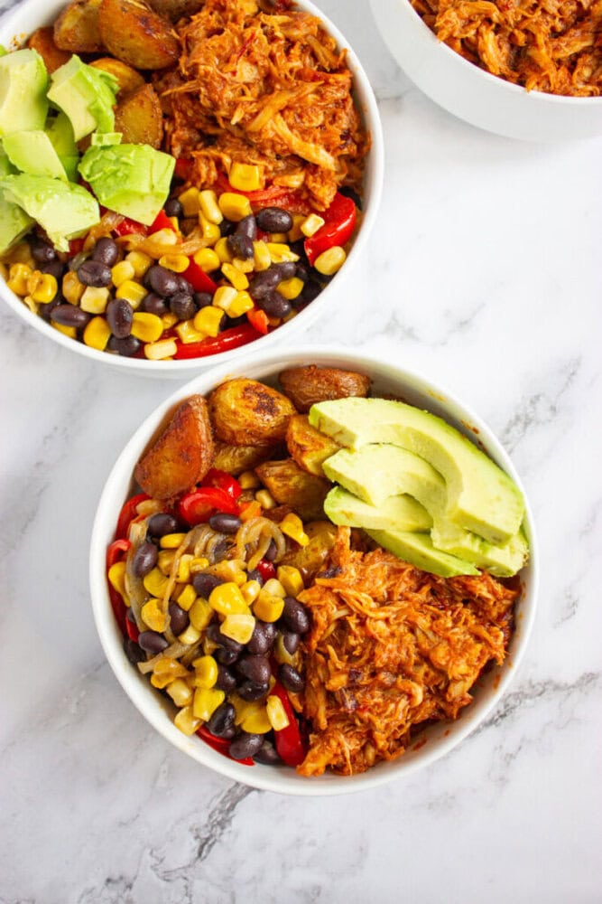 Two white bowls filled with shredded chicken, black beans, corn, roasted potatoes, red bell peppers, and sliced avocado, placed on a white marble surface.