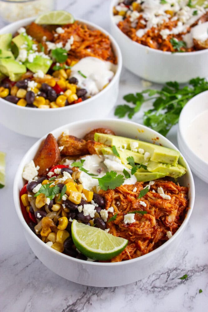 A bowl filled with shredded chicken, black beans, corn, sliced avocado, lime, potatoes, and a drizzle of white sauce, garnished with fresh cilantro.