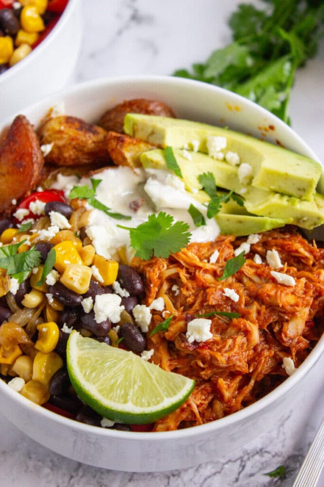 A burrito bowl with shredded chicken, black beans, corn, roasted potatoes, avocado slices, lime wedge, sour cream, feta cheese, and cilantro.