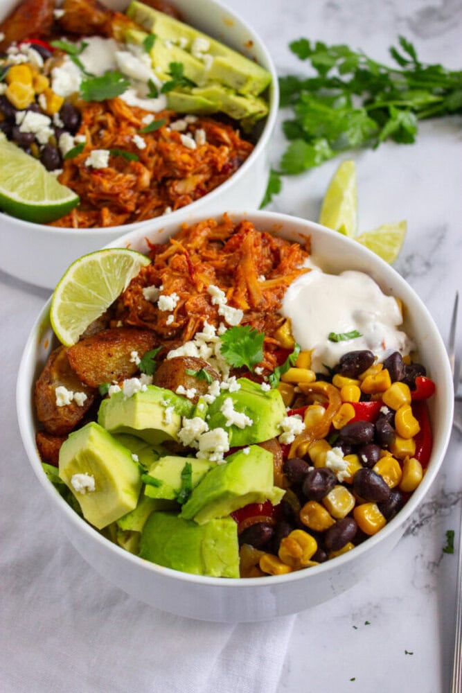 A bowl filled with shredded chicken, avocado slices, black beans, corn, diced potatoes, crumbled cheese, a lime wedge, and a drizzle of white sauce, garnished with cilantro.