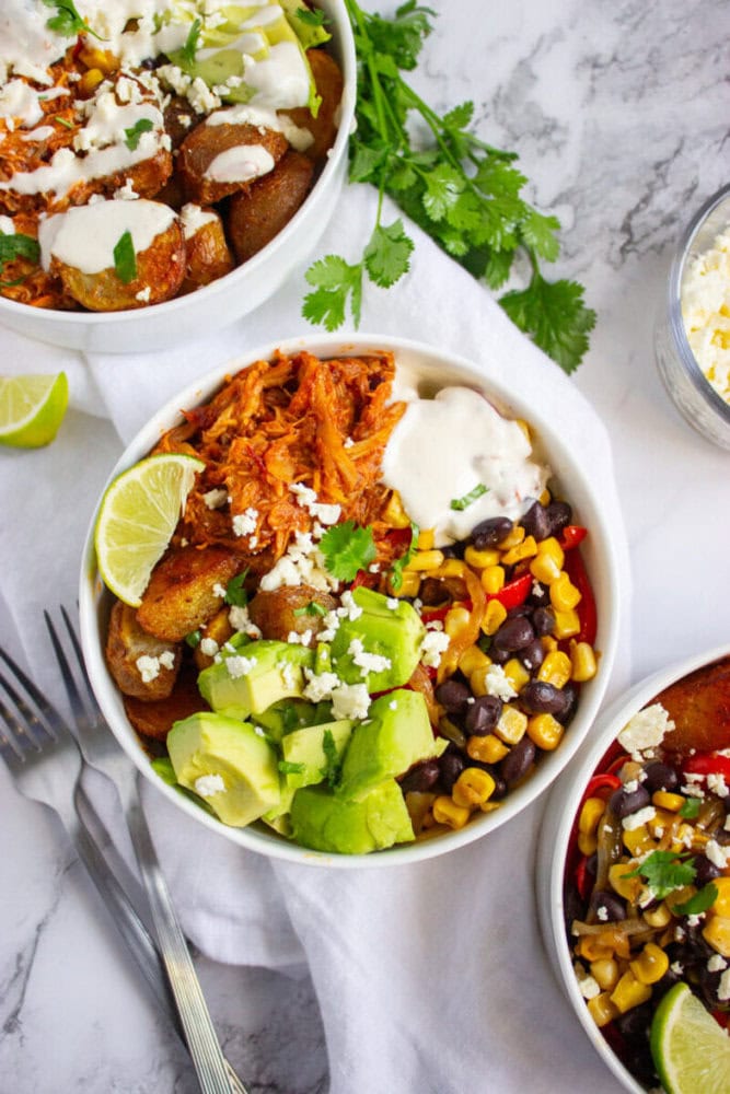 A bowl with shredded chicken, black beans, corn, avocado, roasted potatoes, cheese, and a dollop of white sauce, garnished with cilantro and a lime wedge, on a marble surface.
